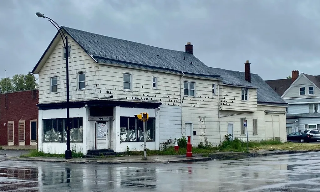 Derelict white former shop on a wet street corner with obscured windows and tall weeds.