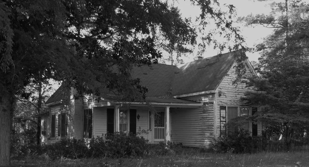 Deteriorated old wooden house among trees, in black and white.