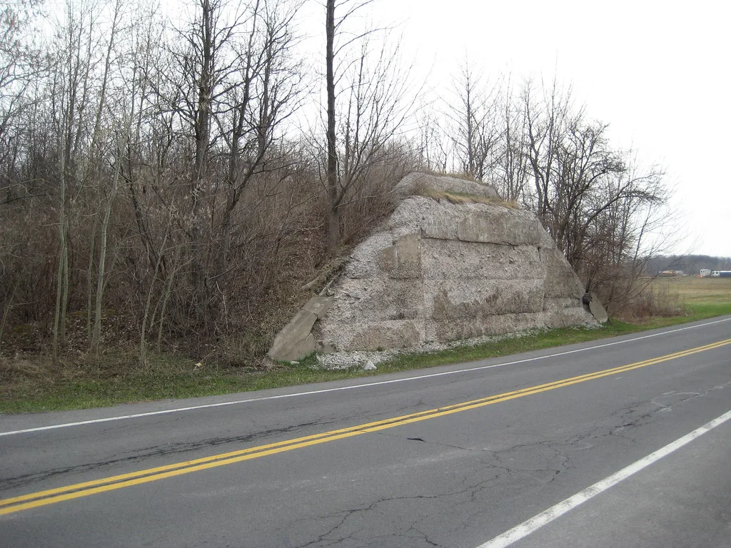 Abandoned old bridge abutment of eroded concrete beside a road in front of bare trees.