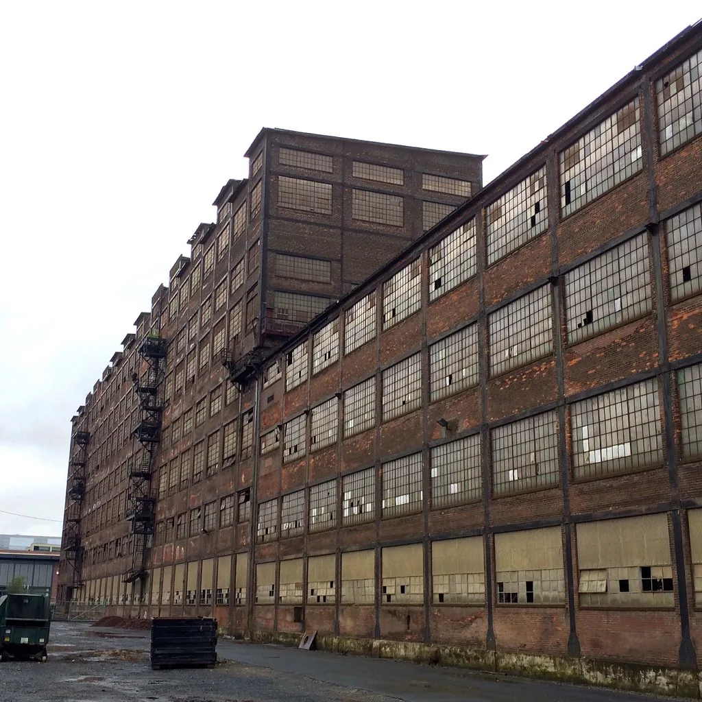 Long multi-storey brick factory with large broken window walls before a deteriorated yard.