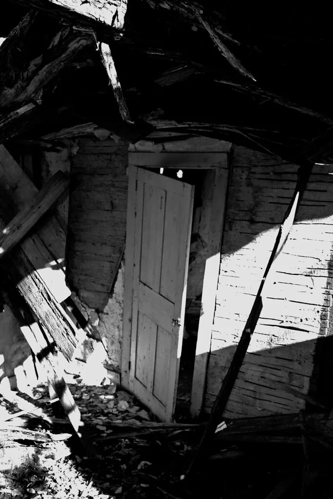 Half-open door beneath broken beams in a ruined wooden interior, in black and white.