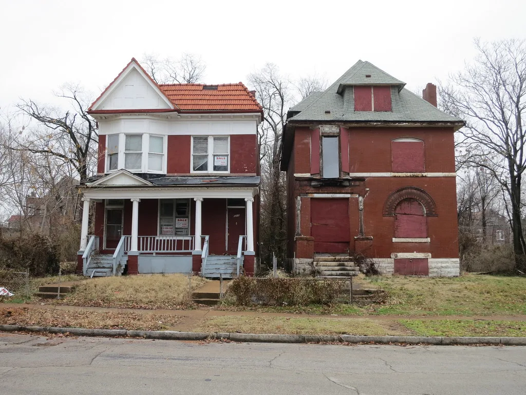 Two red houses side by side, the right-hand one abandoned with boarded openings and holes in its roof.