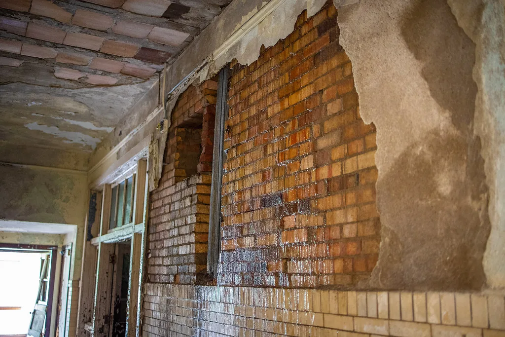 Abandoned corridor with brickwork exposed by fallen plaster, flaking door frames and a deteriorated ceiling.
