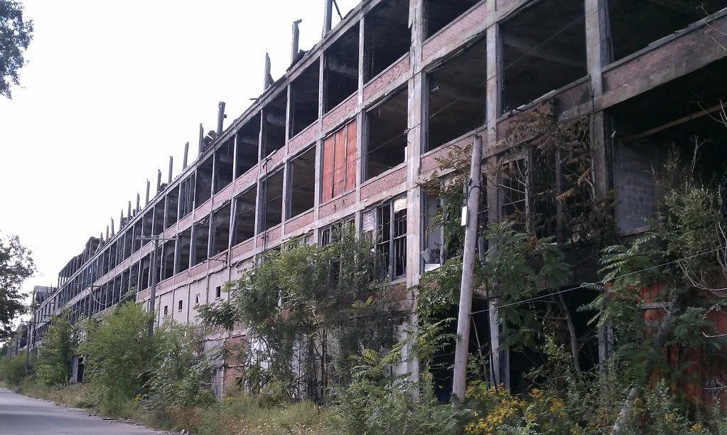 Multi-storey abandoned factory facade with large empty window bays and encroaching shrubs beside a road.
