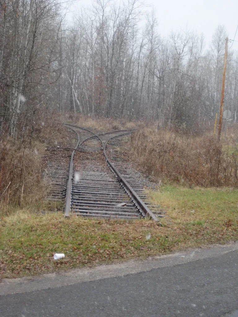 Ancien aiguillage rouillé dont les rails s’arrêtent dans l’herbe au bord d’une route.