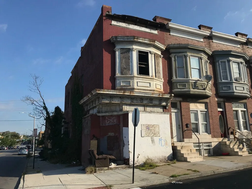 Abandoned red-brick corner house with a broken bay window, sealed storefront and an armchair left by the entrance.