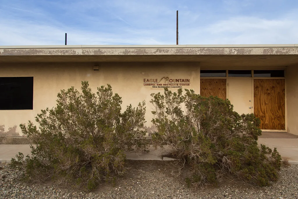 Low beige facade with glassless or boarded openings behind desert shrubs.