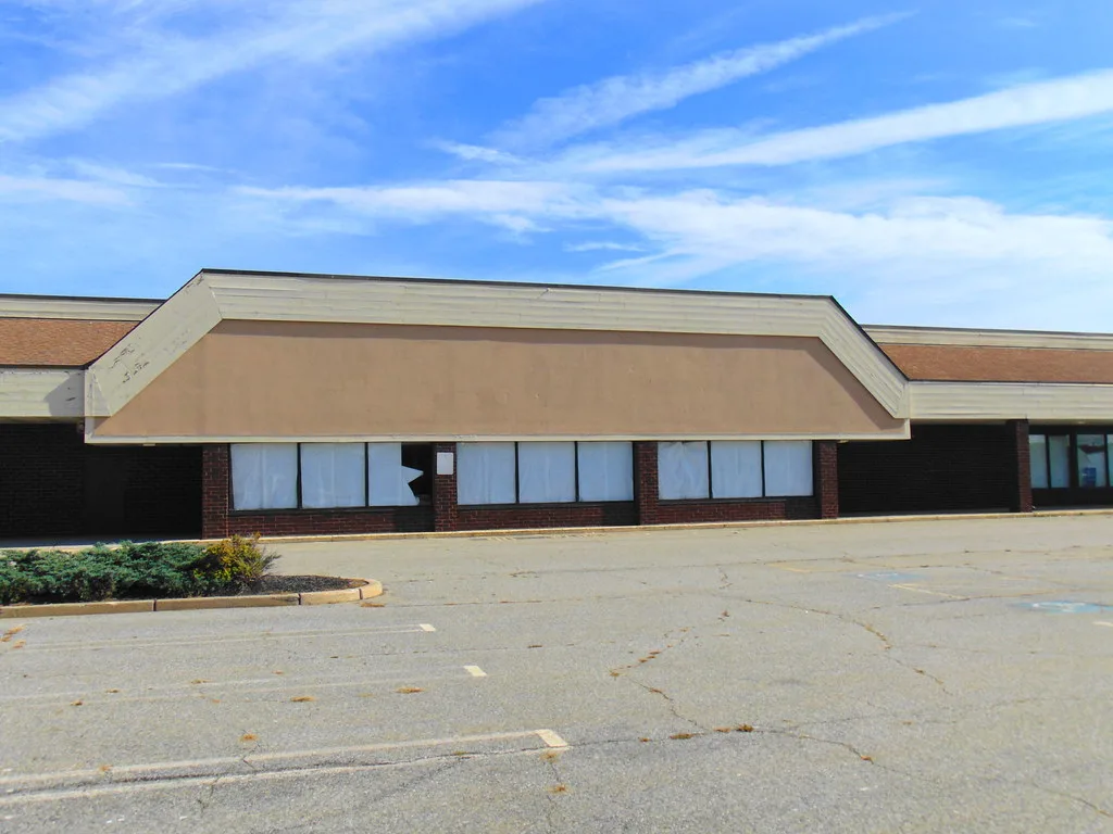 Empty former shop facade with a broken window before a cracked parking lot sprouting weeds.
