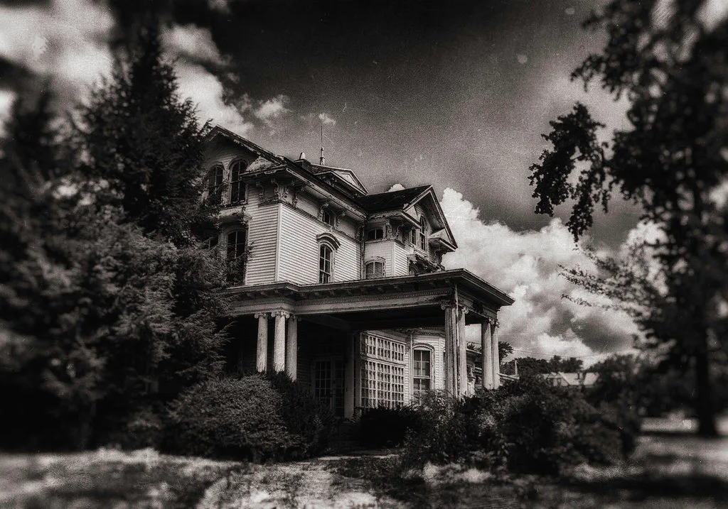 Black-and-white view of a large abandoned house with a columned porch, empty upper windows and encroaching bushes.