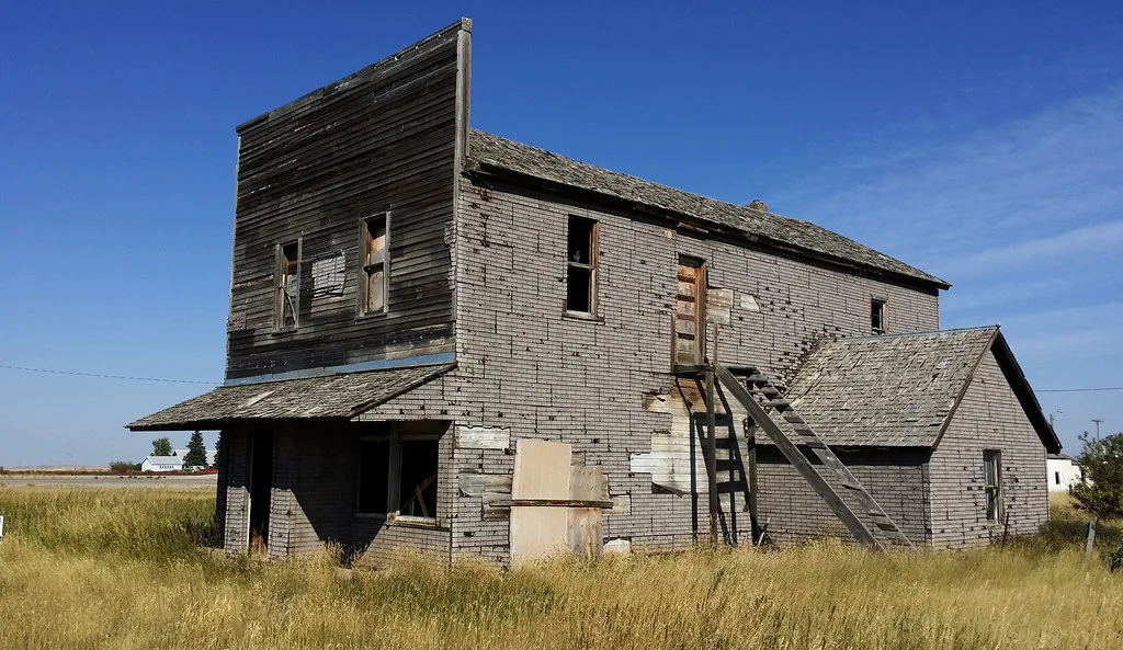 Old building with empty windows and an exterior staircase amid tall grass.