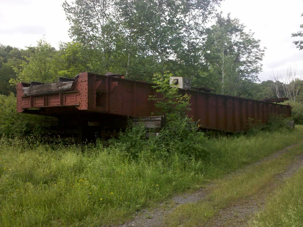 Abandoned former railway bridge deck with rusty girders resting among grass and shrubs.