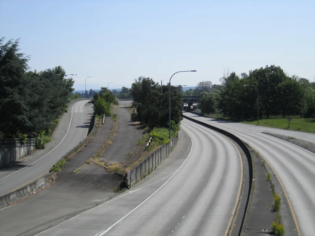 Disused road ramp with cracked asphalt overgrown by trees between two roads.