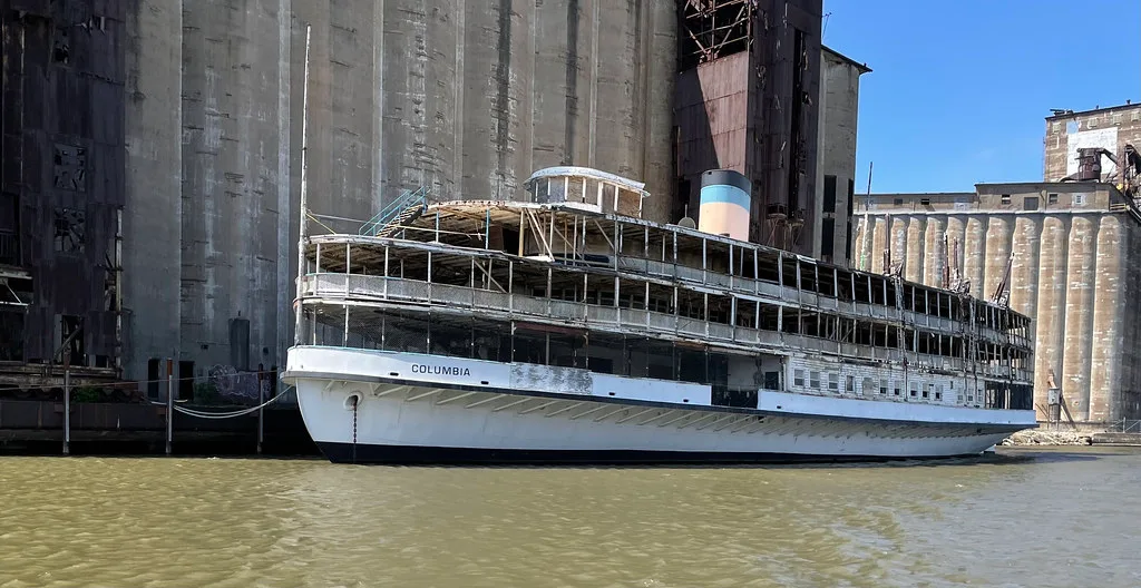 Old white boat with deteriorated decks in front of large concrete silos.