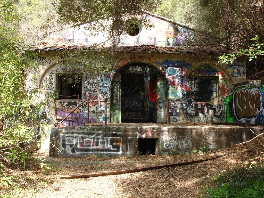 Small abandoned house covered in graffiti, with an arched entrance and empty windows beneath branches.