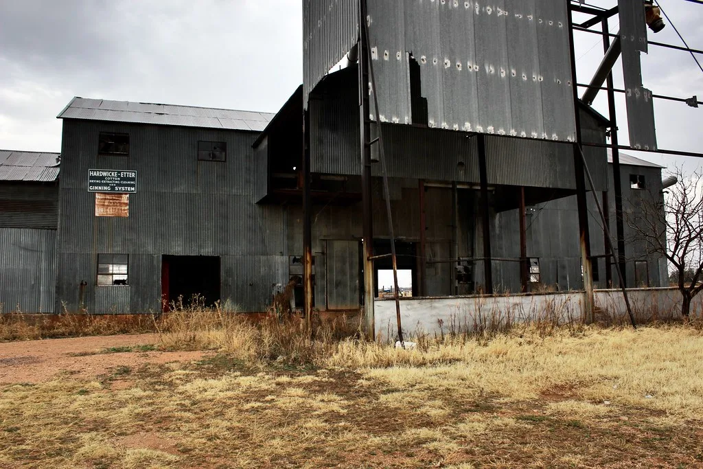 Abandoned former sheet-metal industrial installation with missing cladding and broken windows above dry grass.