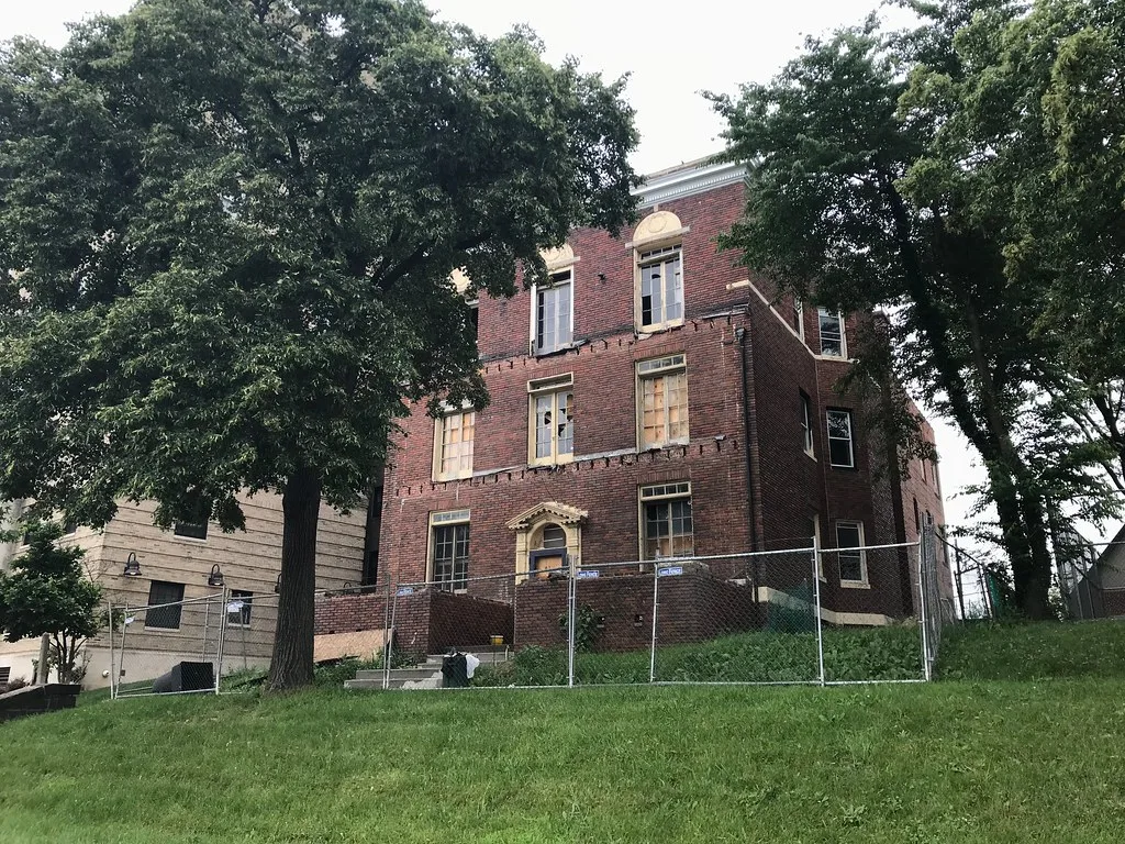 Old brick building with broken windows behind a fence.