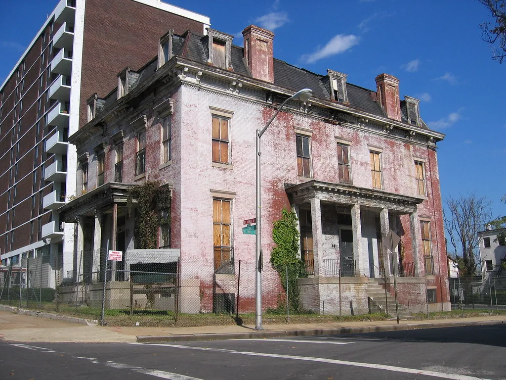 Large abandoned pink-and-white house with wood-covered windows and columned porches overgrown with vines.