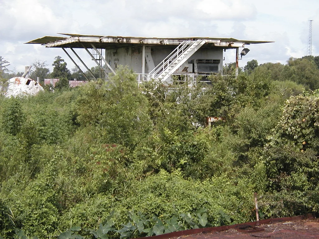 Abandoned flat-roofed utility building with exterior stairs above dense scrub.