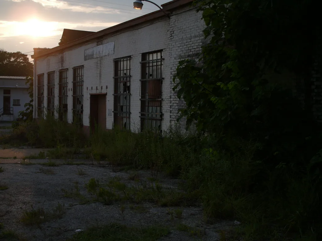 White brick workshop with tall broken windows and an overgrown yard at sunset.