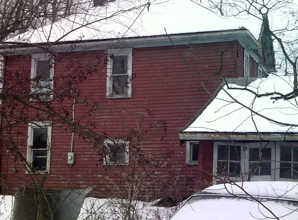 Maison rouge délabrée aux fenêtres brisées derrière des branches, dans la neige.
