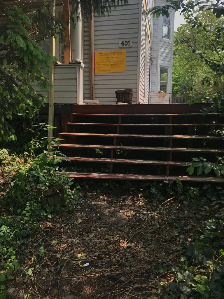 Overgrown exterior staircase leading to a boarded old house.