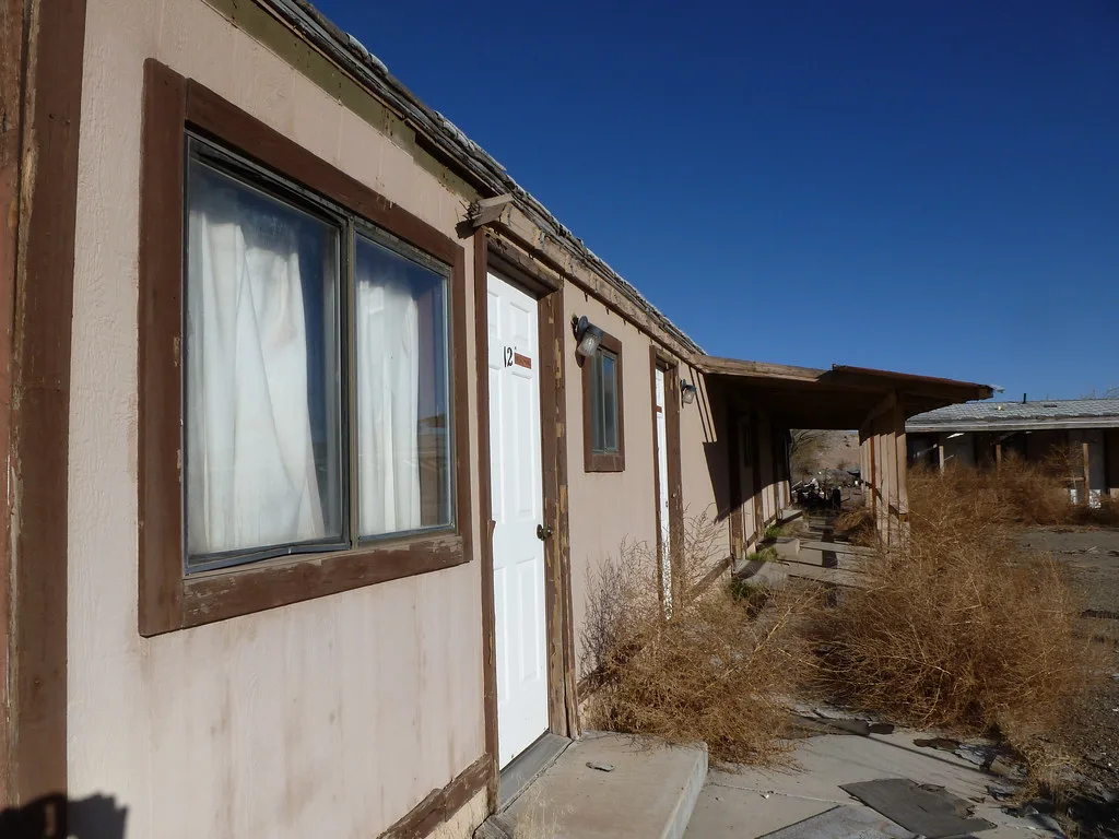 Row of abandoned motel rooms with scrub-choked entrances, a deteriorated canopy and debris along the walkway.