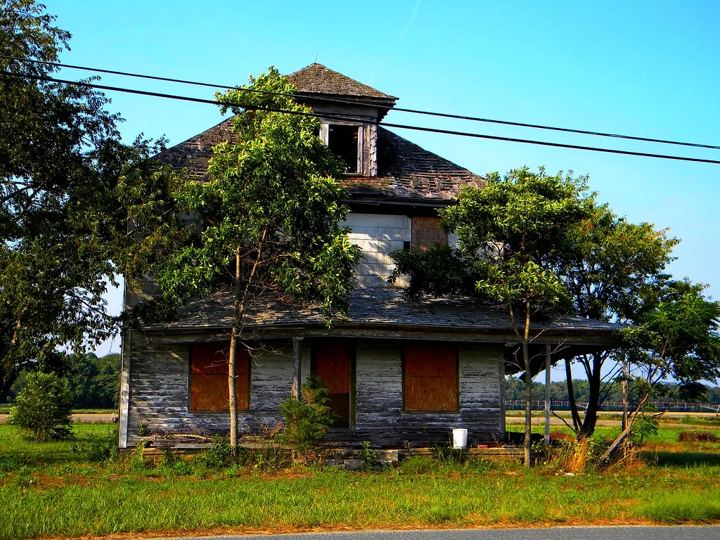 Abandoned wooden house with boarded windows and an empty dormer behind trees.