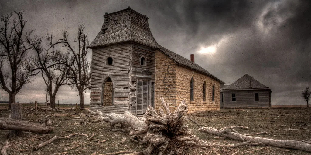Église abandonnée de pierre et bois à clocher tronqué, devant des troncs couchés sous un ciel sombre.