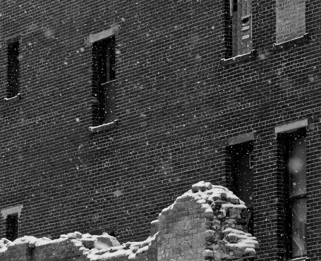 Collapsed brick wall in front of an abandoned facade with empty windows in snow.