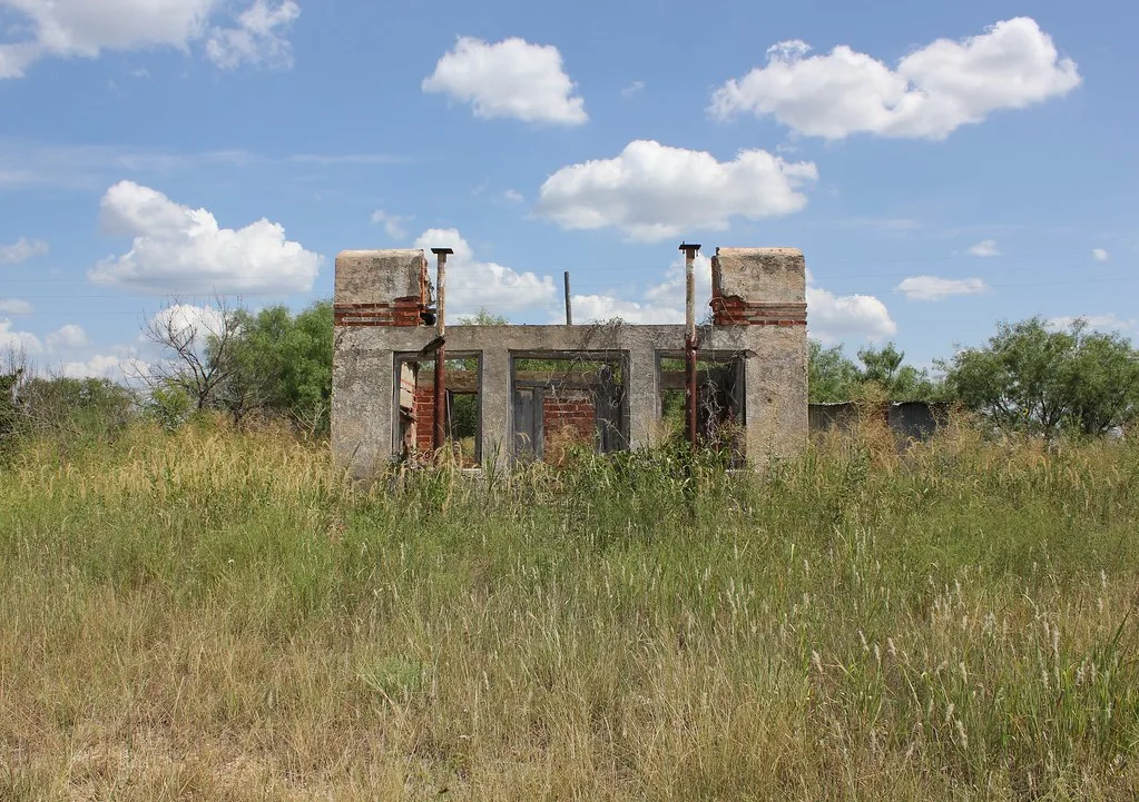 Remains of a small roofless petrol station in tall grass.