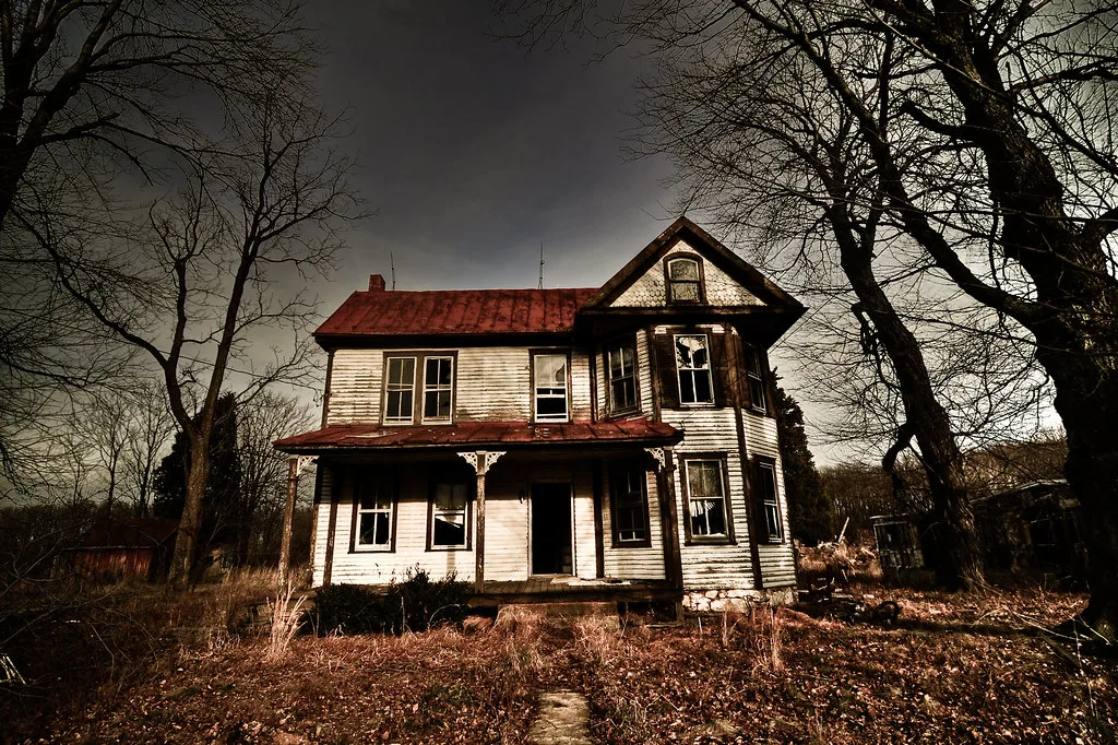 White abandoned house with a bay window and rusty red roof, with broken windows among leafless trees.