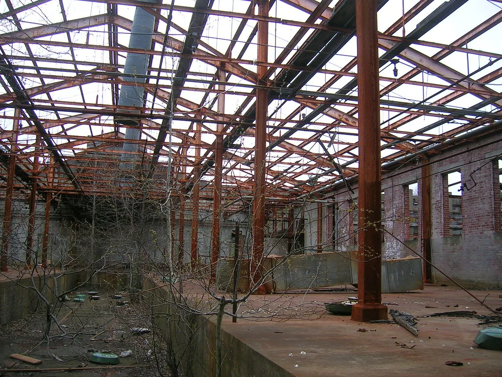 Roofless abandoned industrial hall with rusty steel framing and brick walls, with trees growing in floor pits.
