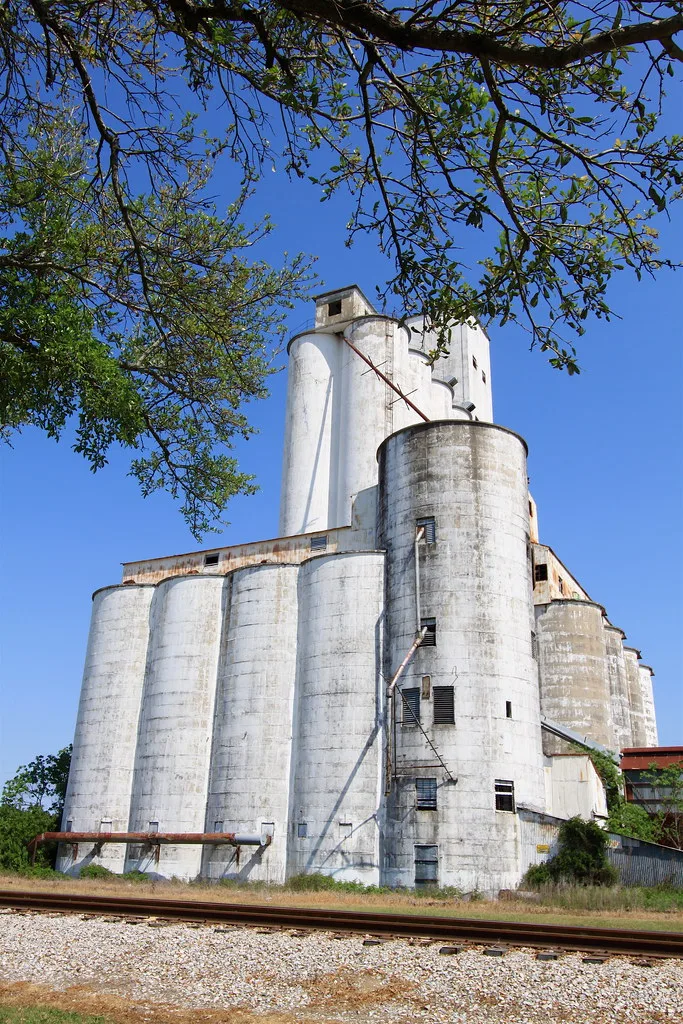 Old white concrete silos with deteriorated openings beside a railway.