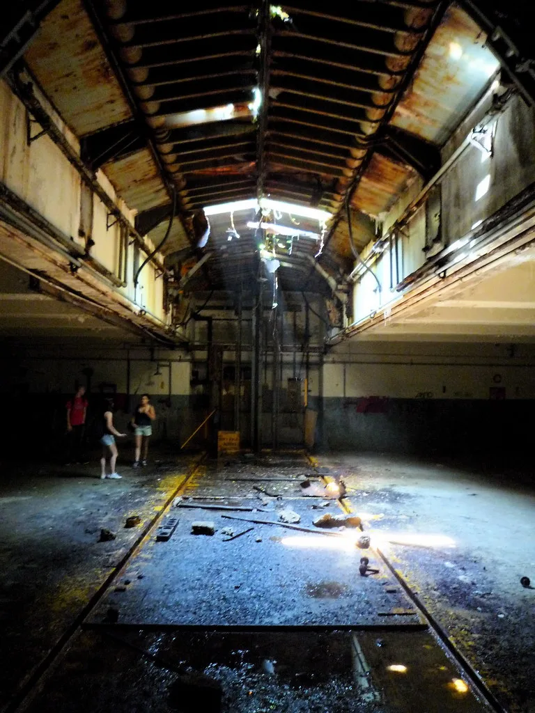 Abandoned hall with a broken roof and peeling walls, with floor rails, debris and puddles beneath light entering through the roof.