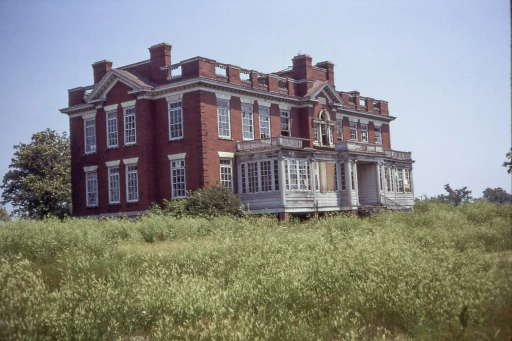 Grande maison abandonnée de briques rouges à vérandas blanches et fenêtres brisées dans de hautes herbes.