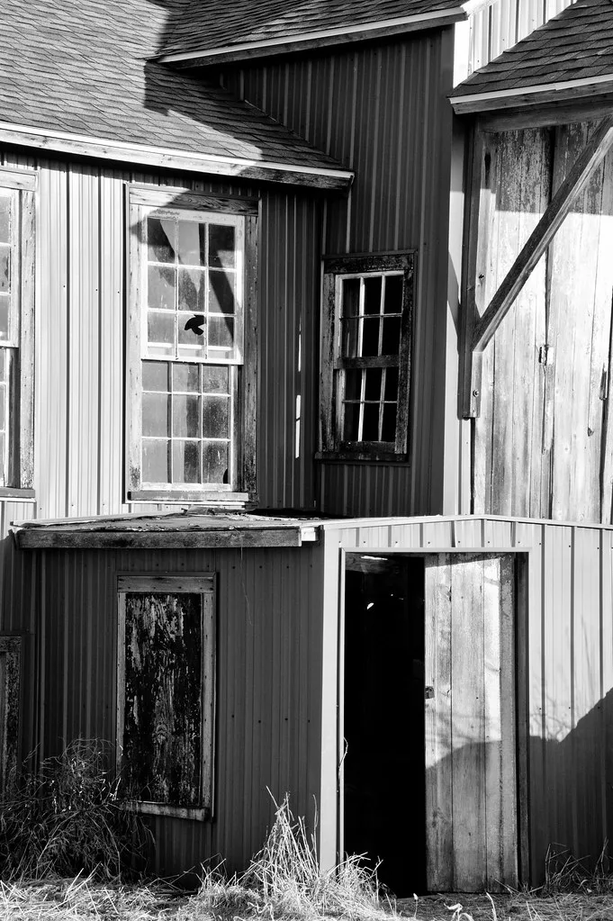Old facade with broken windows and an open doorway amid weeds.