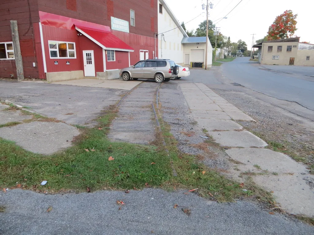 Ancien tronçon de rails dans le béton devant un bâtiment rouge et des voitures garées.