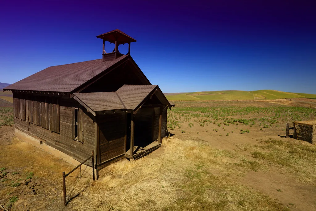 Old wooden building with a bell turret, boarded windows and a deteriorated porch in a dry field.