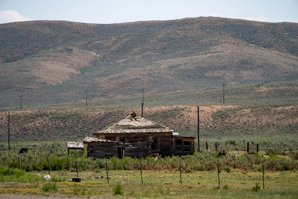 Ruined wooden house below dry hills, with a damaged roof and empty windows.