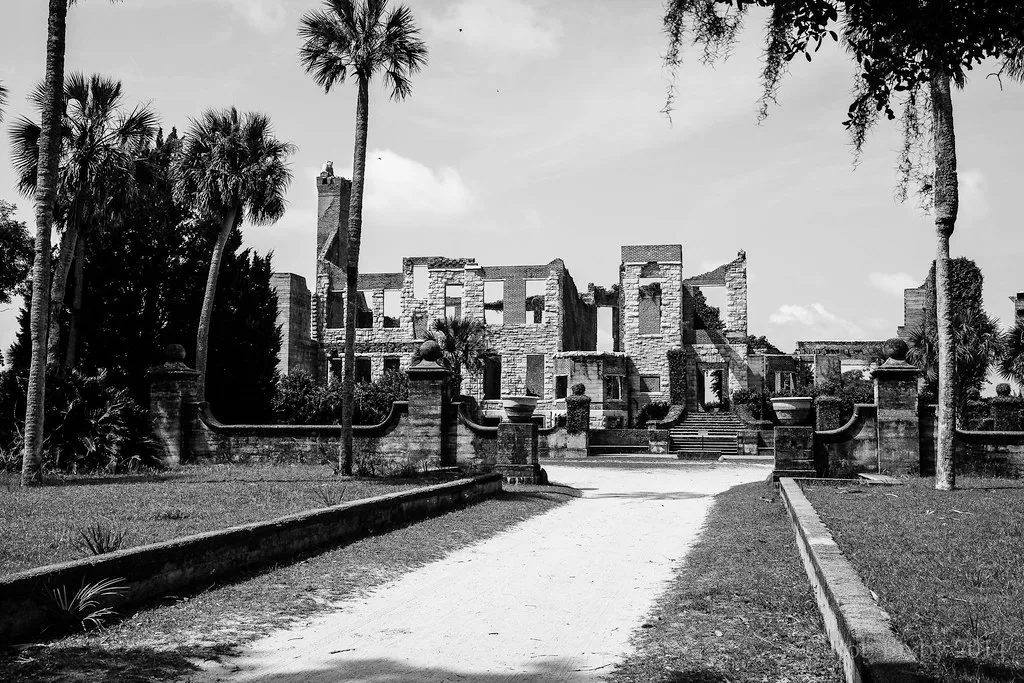 Ruined roofless stone mansion facade at the end of a palm-lined path.