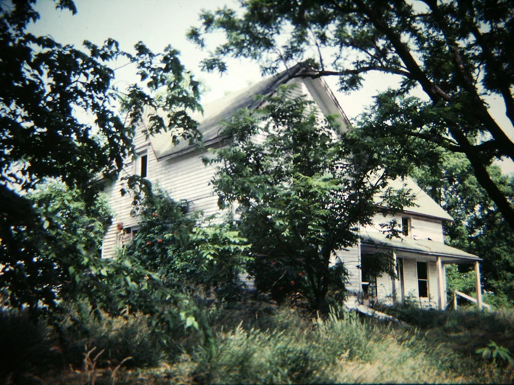 Large abandoned white-clad house with a deteriorated porch and empty windows behind trees and tall grass.