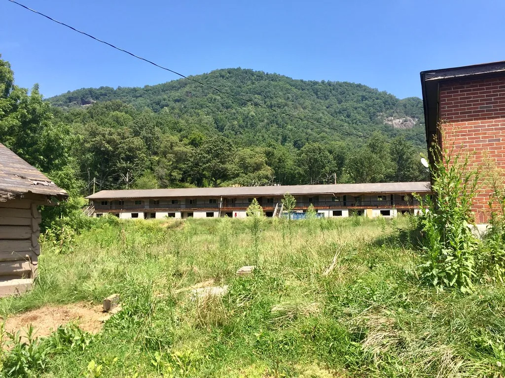 Abandoned old two-storey motel behind an overgrown courtyard at the foot of a wooded hill.