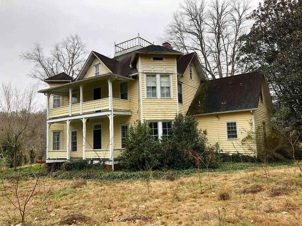 Old yellow house with two verandas surrounded by encroaching vegetation.