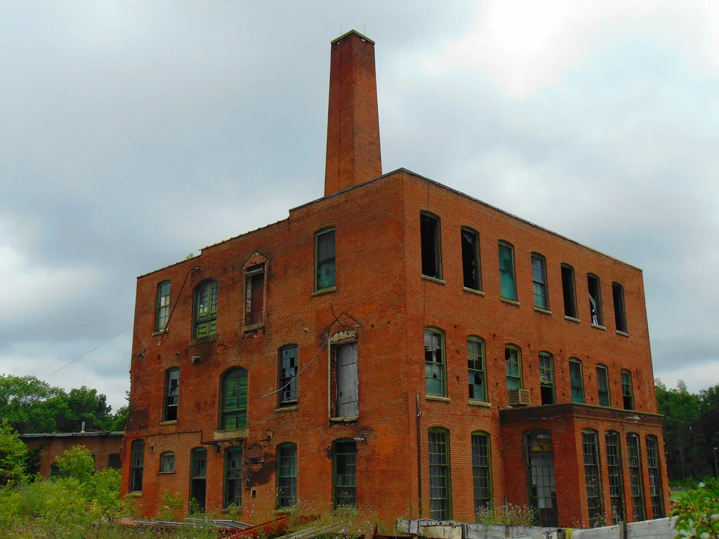 Former red brick factory with broken windows and a tall chimney.