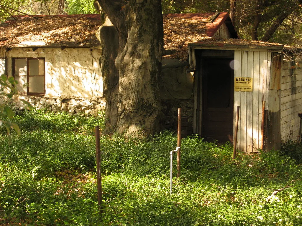Small building with cracked render and a worn wooden porch beneath a leaf-covered roof.