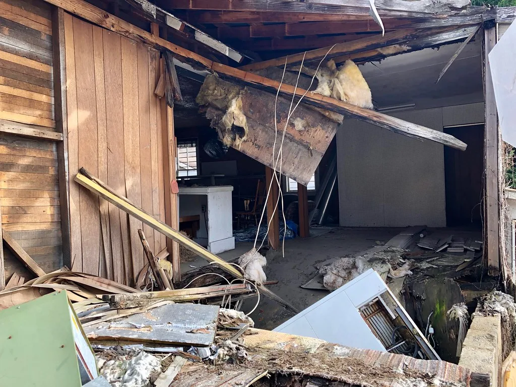 Abandoned room exposed by a collapsed wall, with broken beams, hanging insulation and furniture debris.