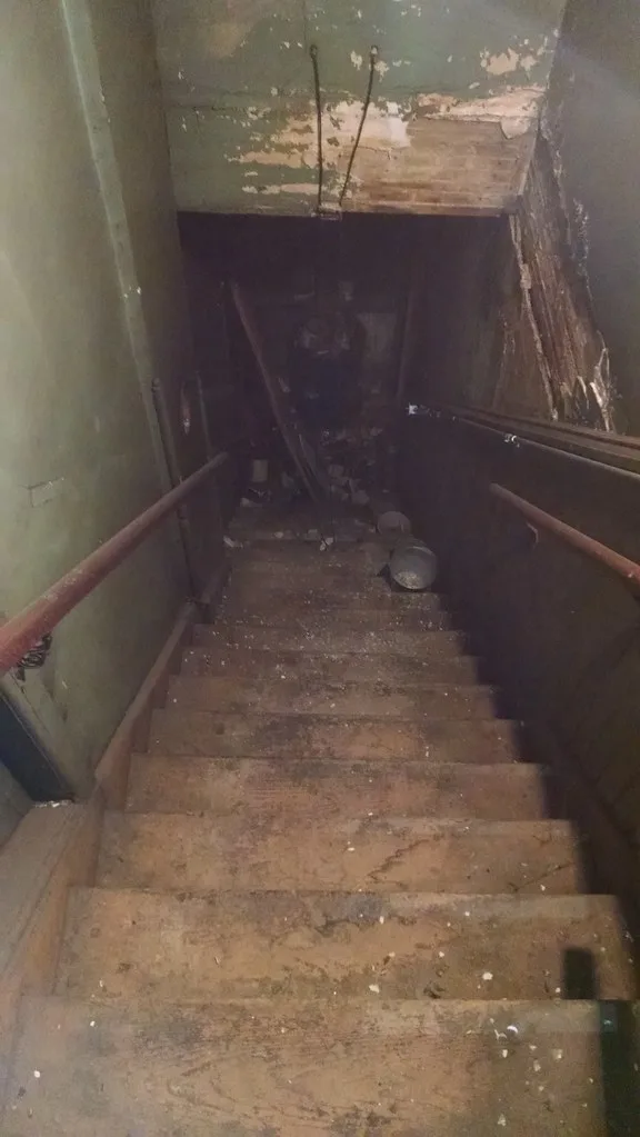 Abandoned indoor stairway with peeling green walls, rusty handrails and debris at the bottom.
