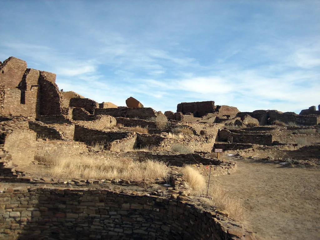 Extensive roofless ochre stone ruins with stepped walls and dry grass beside a dirt path.