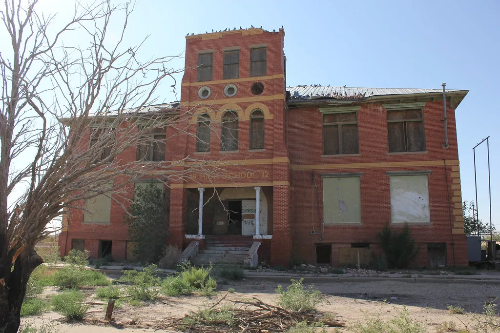 Abandoned red-brick school with a central tower, sealed openings and a damaged roof covered with pigeons.