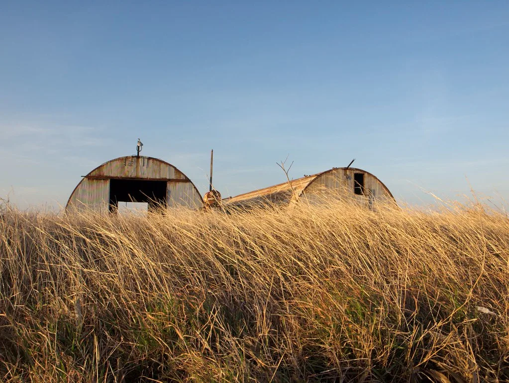 Two old rusty round-roofed sheds behind tall dry grass.
